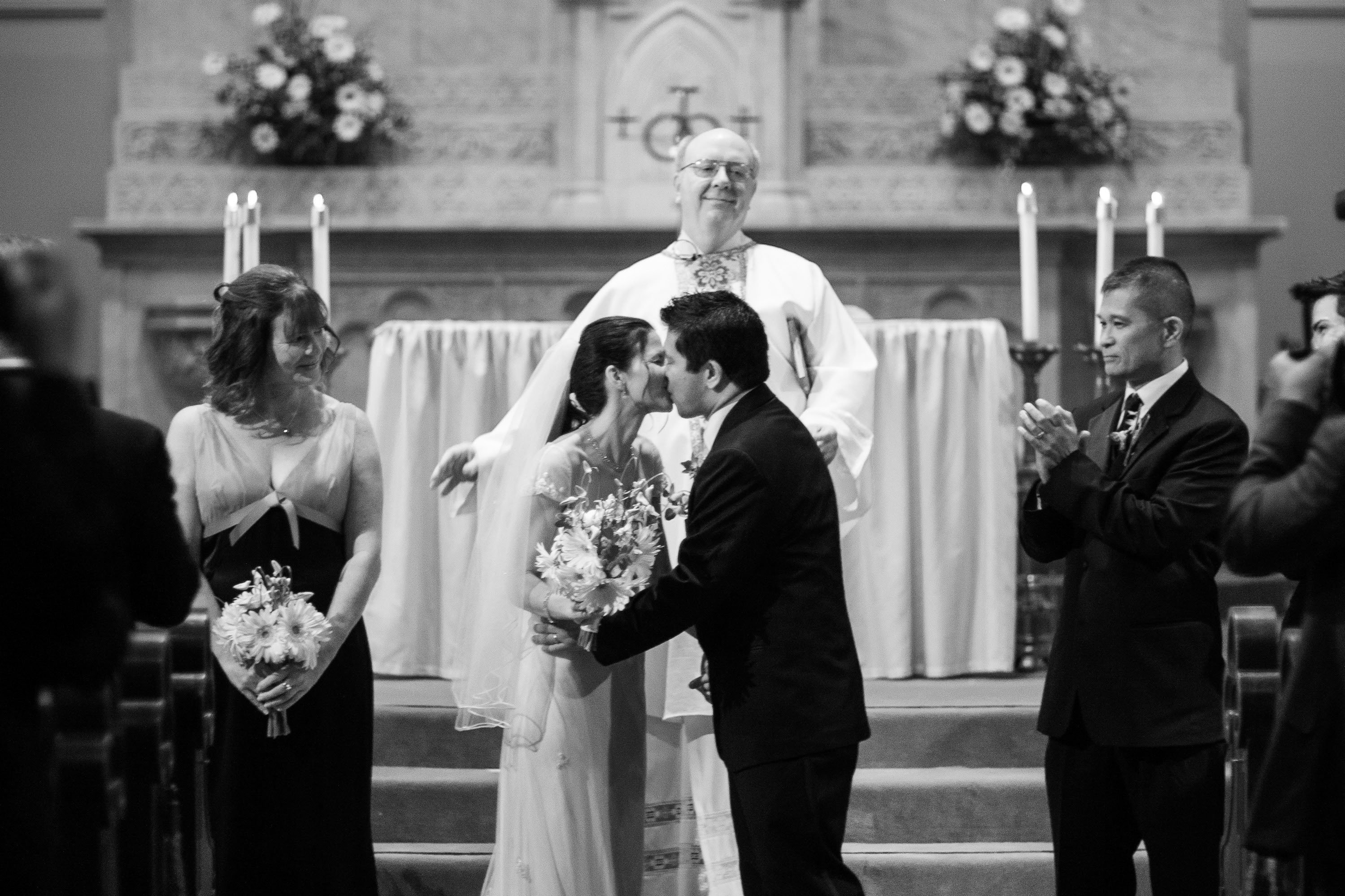 The couple kiss as the bridal party applauds and the pastor looks on with a smile,  at Old St. Mary's Church in San Francisco